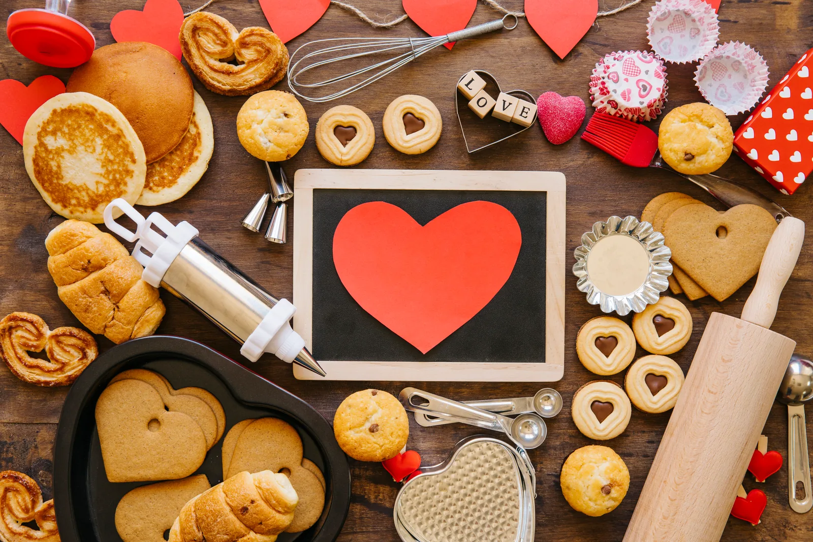 Cakes, biscuits and baking utensils around a rectangular chalkboard topped with a red paper heart.